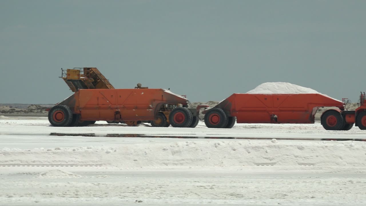 At 0:02, the red hauler continues across the white ground in Guerrero Negro, showing the white material piled above the red container and no visible people.