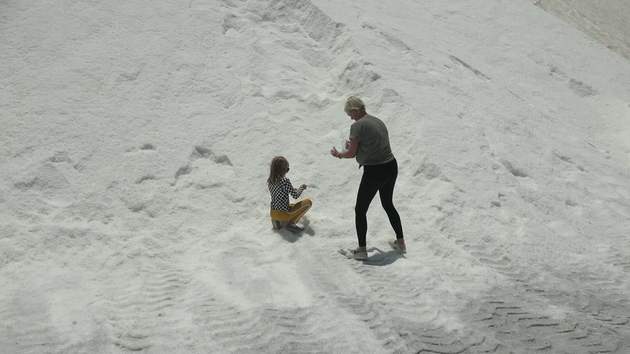 In Guerrero Negro, Mexico, a child sits on pale textured sand while an adult stands nearby with hands raised slightly at the start of the clip.