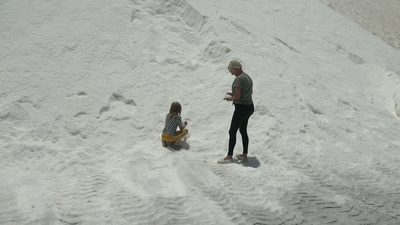In Guerrero Negro, Mexico, the child remains seated with yellow pants visible while the adult looks downward with hands near the body on the open sand.