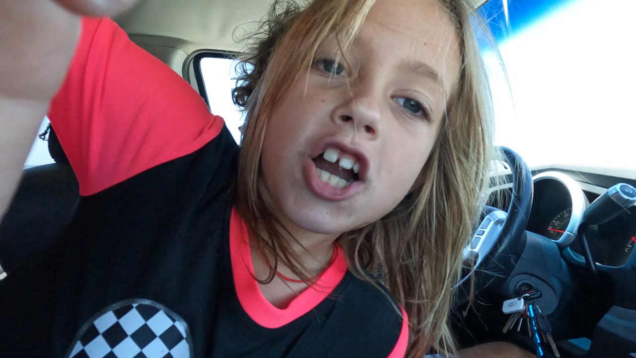 In Baja California, Mexico, a child in a black and red checkered shirt leans close to the camera inside a car with their mouth open and the steering wheel visible behind them.