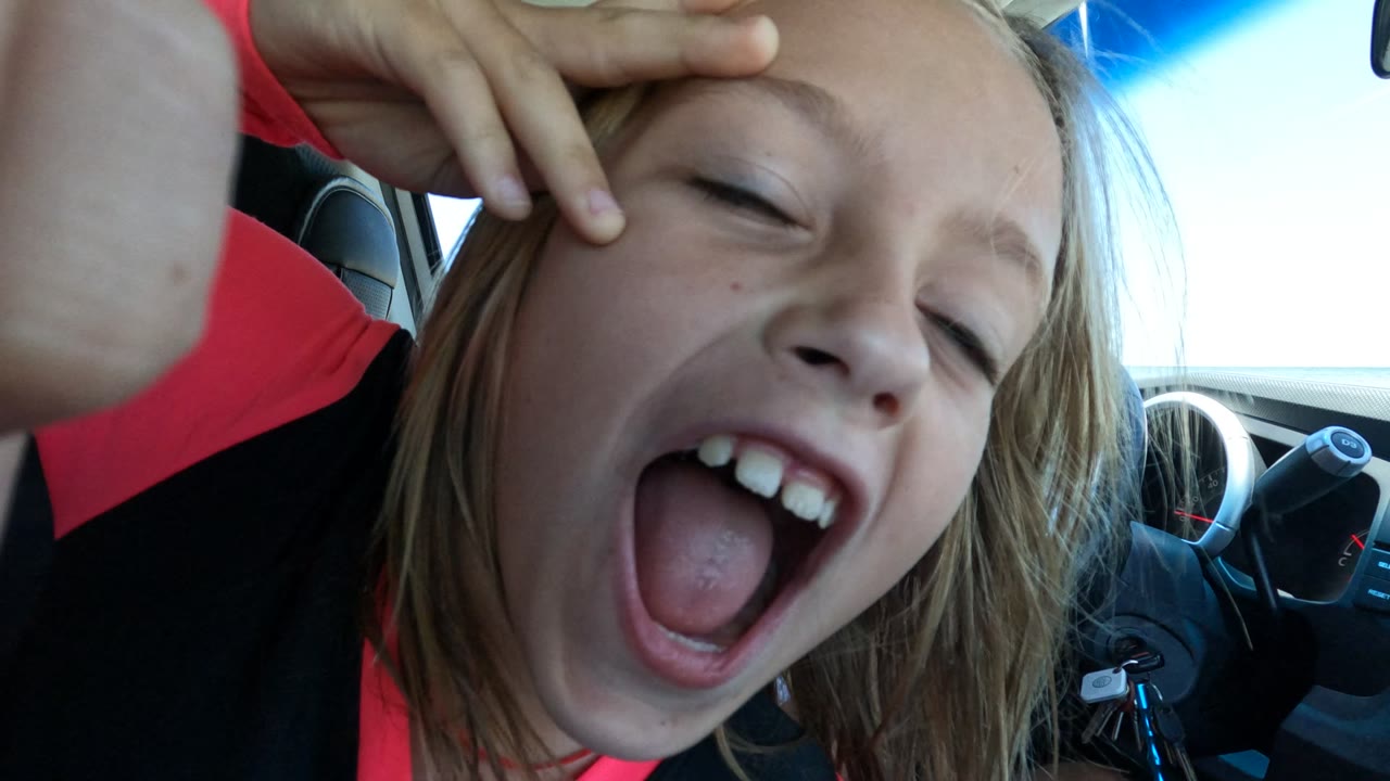 In Baja California, Mexico, the child winks while holding fingers near their forehead, with the car steering wheel, dashboard, and window light behind them.