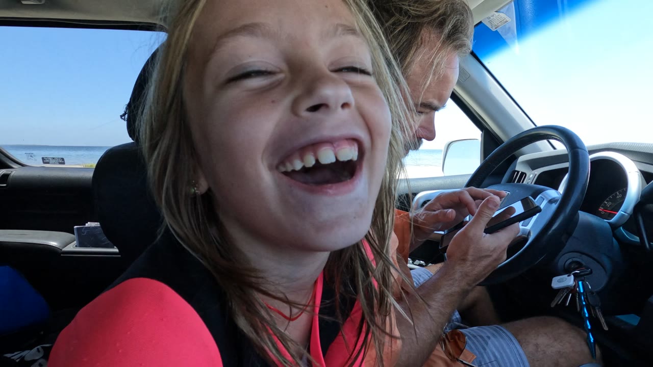 In Baja California, Mexico, a child laughs close to the camera inside a car while a driver looks down near the steering wheel and the ocean is visible through the window.