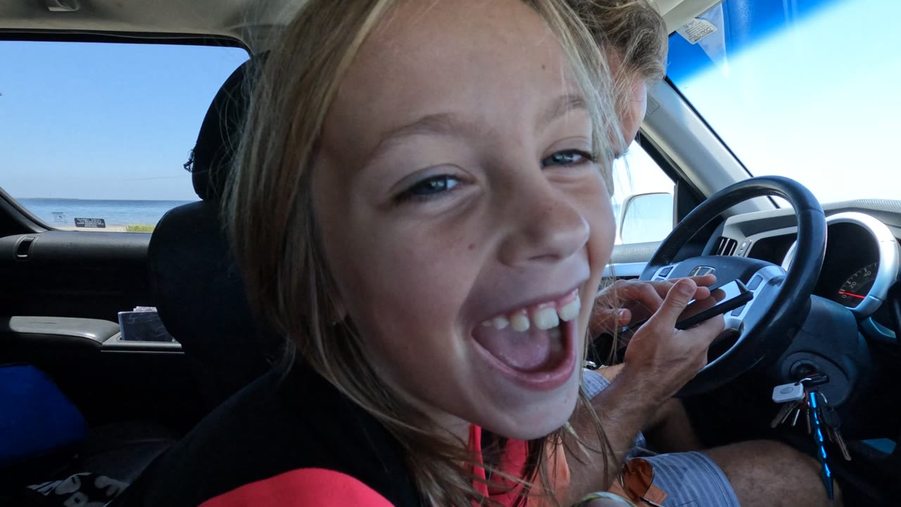 Child grinning with one eye partly closed inside a car near the ocean.
