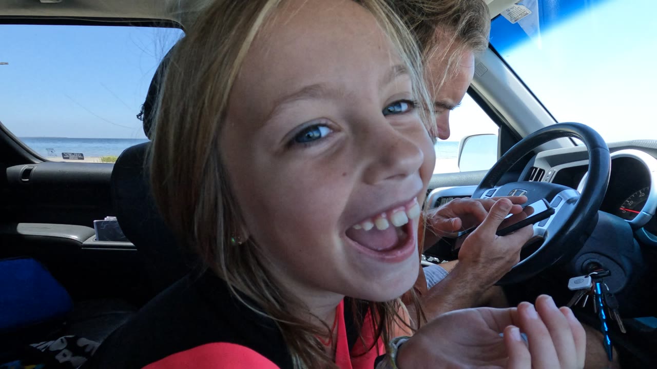 In Baja California, Mexico, the child smiles widely and raises a hand near her face while the driver sits behind the wheel and bright ocean light enters through the window.