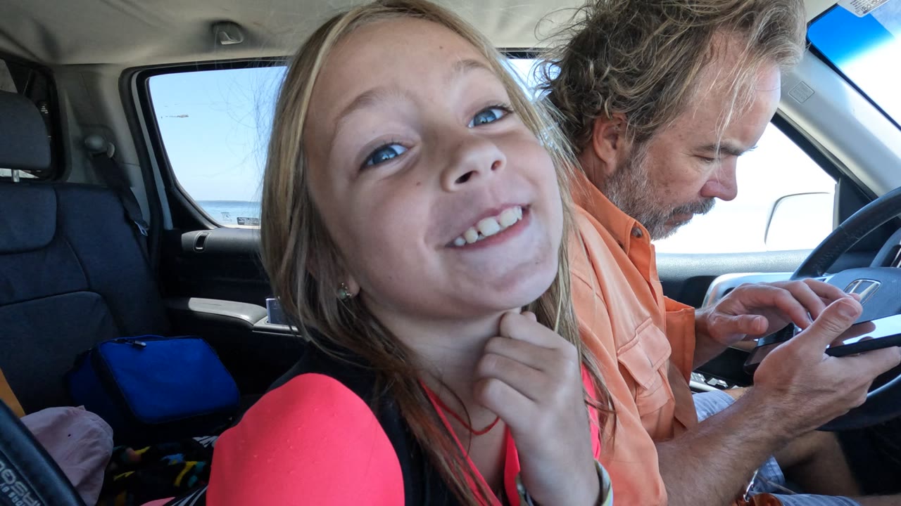 At 0:00 in Baja California, Mexico, a young girl smiles from the front passenger area while an adult driver in an orange shirt looks down at a phone near the steering wheel.