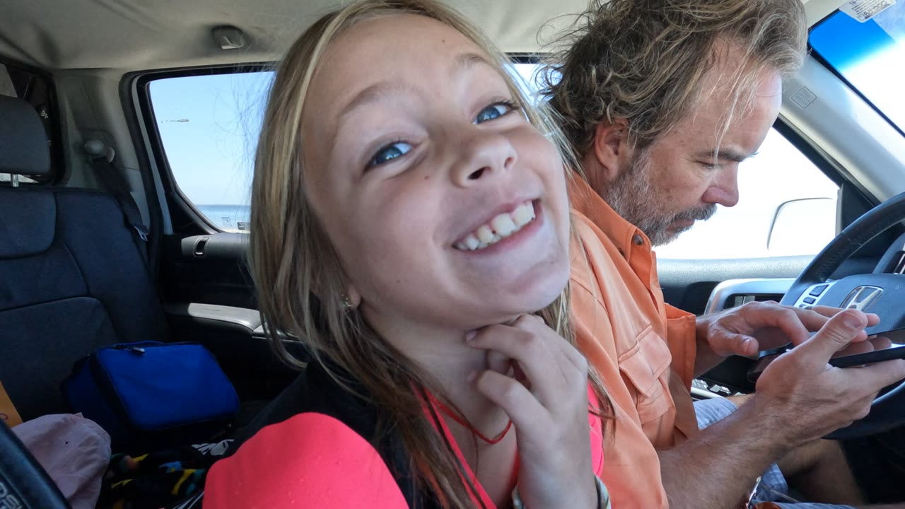 Girl holds her chin and smiles as the driver looks down at a phone.