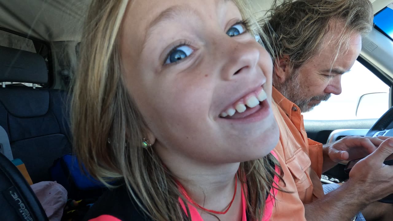 In Baja California, Mexico, a blonde girl smiles close to the passenger-side camera while an adult male driver looks down at a phone near the steering wheel.