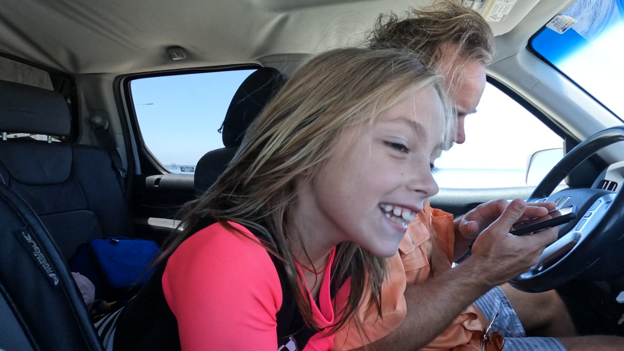 Girl leans forward smiling as the man holds a phone by the steering wheel.