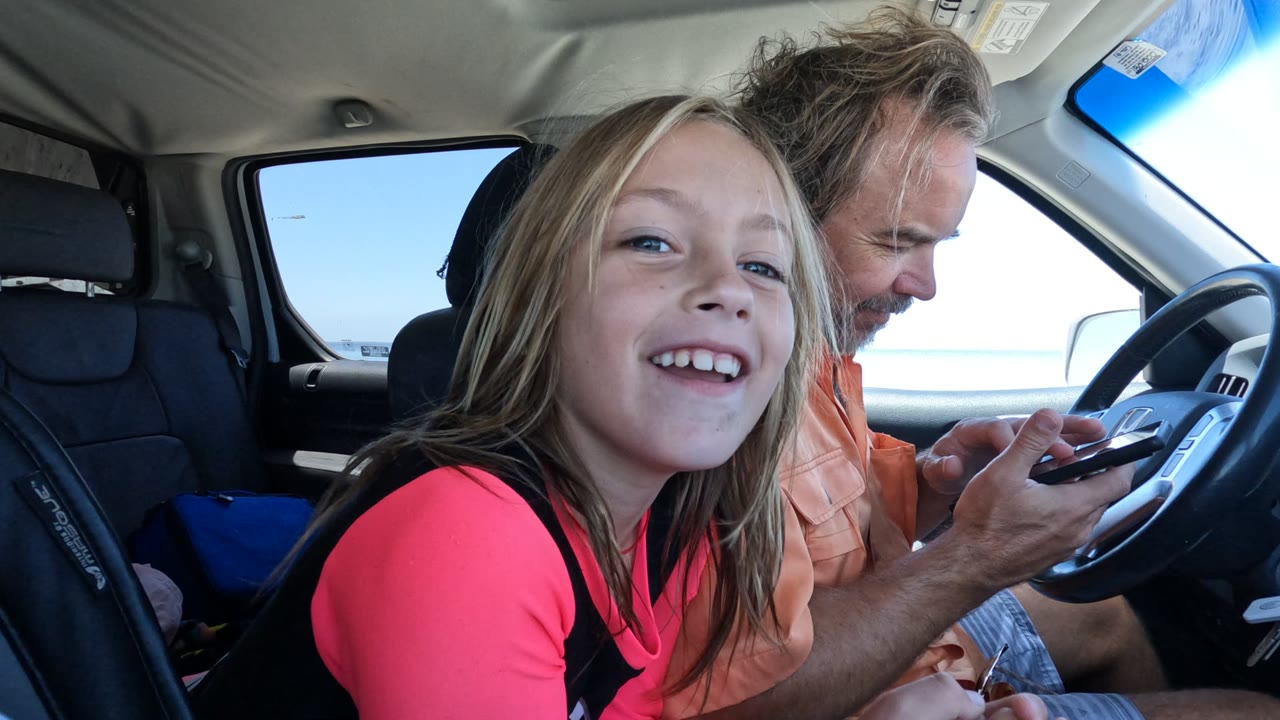 In Baja California, Mexico, the girl turns more toward the camera with a toothy smile as the man behind her looks down at a phone beside the steering wheel.