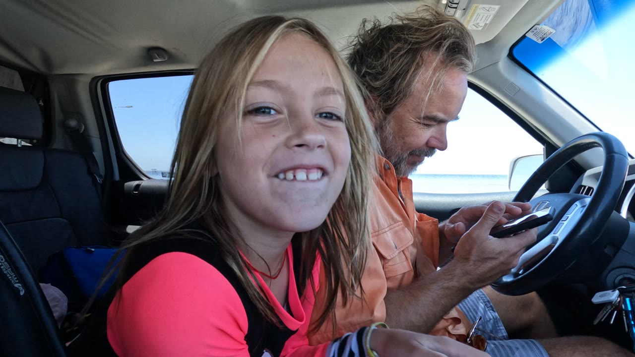 In Baja California, Mexico, a girl in a pink top smiles near the camera from inside a vehicle while a man in an orange shirt looks down at a phone beside the steering wheel.