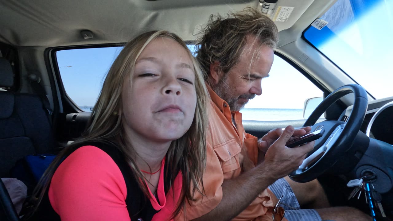 In Baja California, Mexico, the girl leans close to the camera with a squinting expression while the man in the driver's seat continues looking at a phone.
