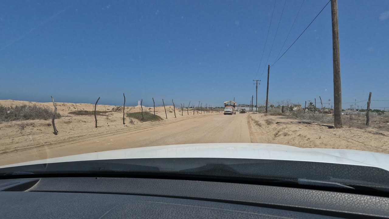 Sandy road seen over a car hood with fence posts and a vehicle approaching.