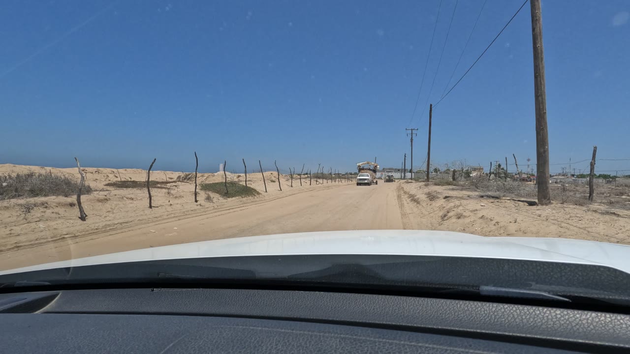 At 0:02, the windshield view shows the sandy Baja California road flanked by dry vegetation and bent fence posts, with an oncoming vehicle slightly larger and no visible people.