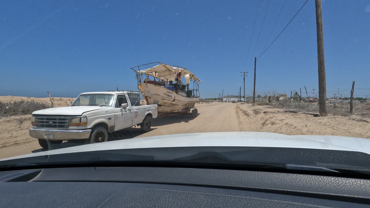 A white pickup towing a weathered boat appears on a sandy Baja California road beside the filming car, with utility poles, fencing, dry scrub, blue sky, and no clearly identifiable people or faces.