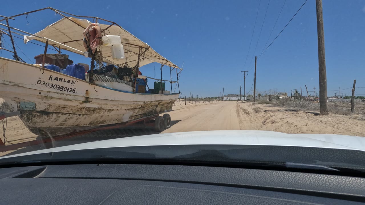 A weathered boat on a trailer fills the right side of the Baja California road view, with registration text on the hull, a canopy, dry fencing, utility poles, open sky, and no clearly identifiable faces.