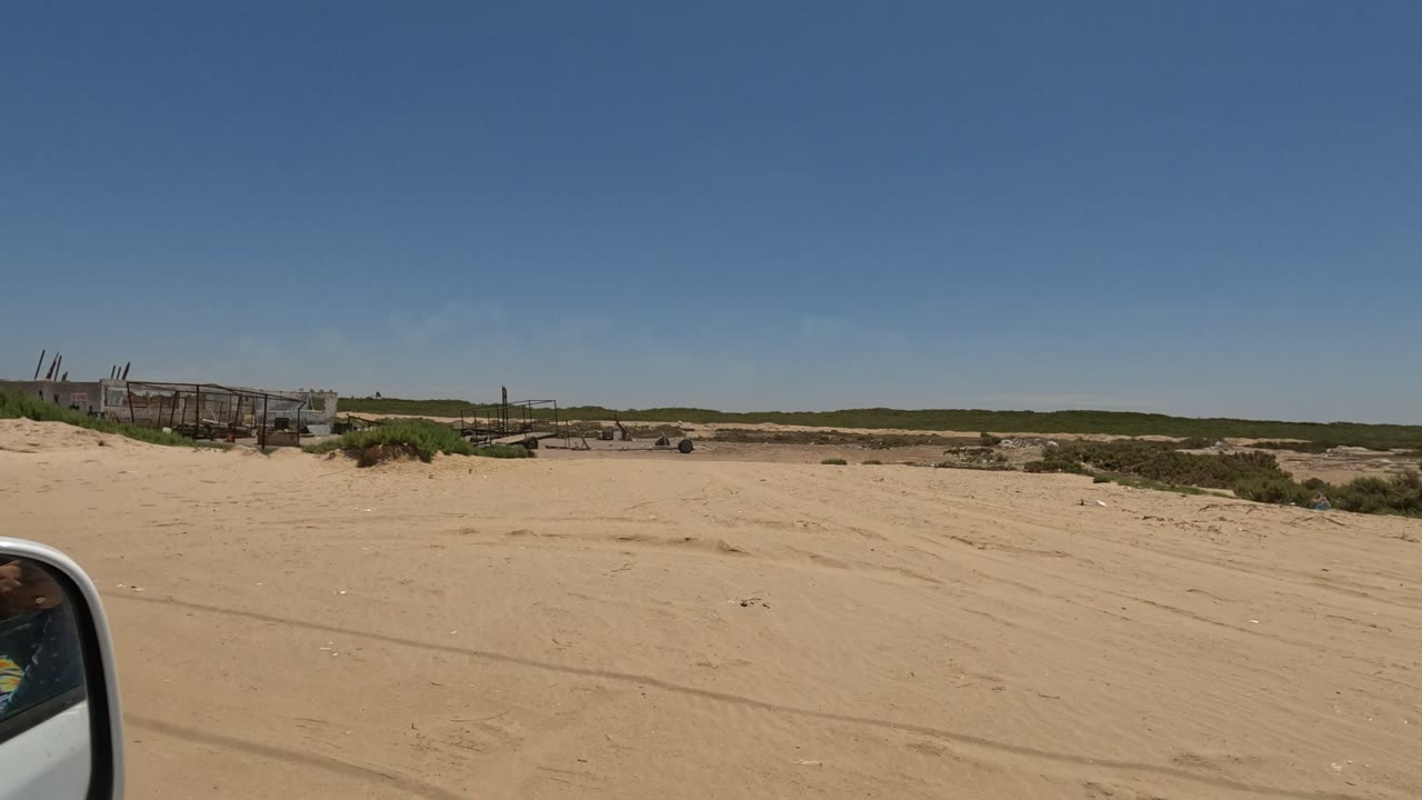 Wide Baja California sandy terrain with low scrub, distant industrial structures, a broad blue sky, and a vehicle mirror at the left edge, with no visible people.