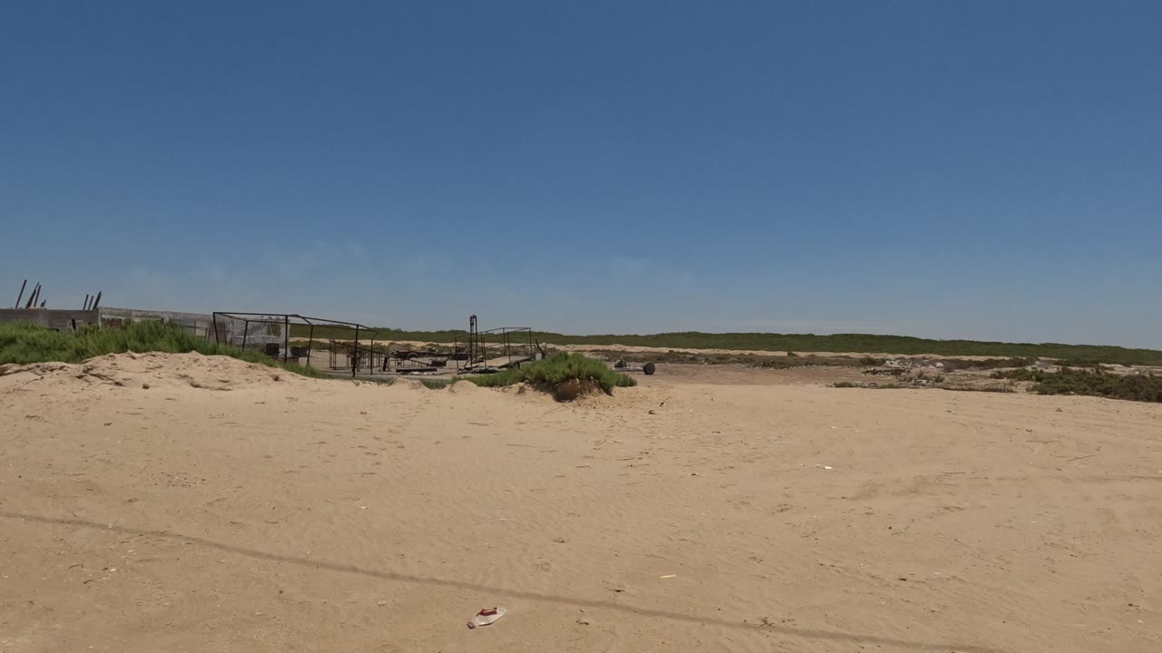 Open sandy area with scrub vegetation, fencing, and low industrial structures in the distance.