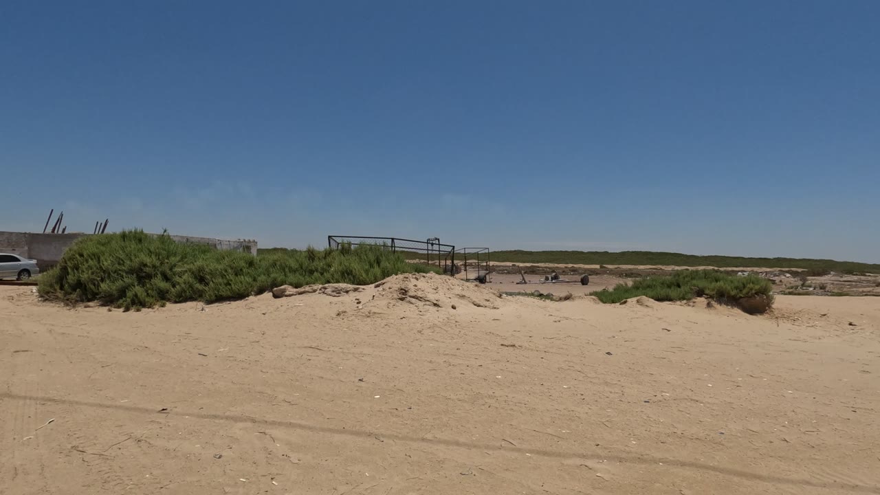 Baja California sandy work area with green shrubs, a white vehicle near the right side, fencing, trailers, and low structures under clear sky, with no visible people.