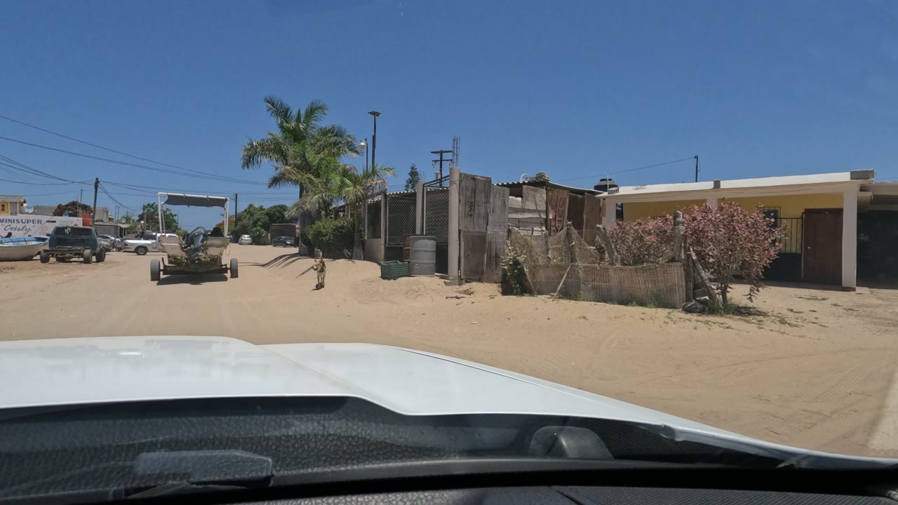 Baja California, Mexico residential roadside seen through the windshield, with a sandy shoulder, yellow house, fencing, palms, parked vehicles, and no visible people.