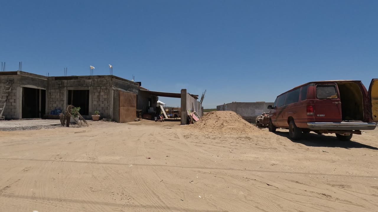 Unfinished cinder block buildings in Baja California stand beside a sandy construction yard with a red van, ladders, sand piles, gravel, and no visible people.