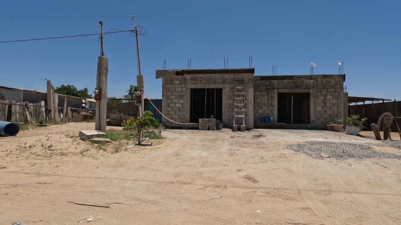 The view shifts along a Baja California sandy road past a utility pole, corrugated and wooden fencing, a blue pipe, sparse vegetation, and no visible people.
