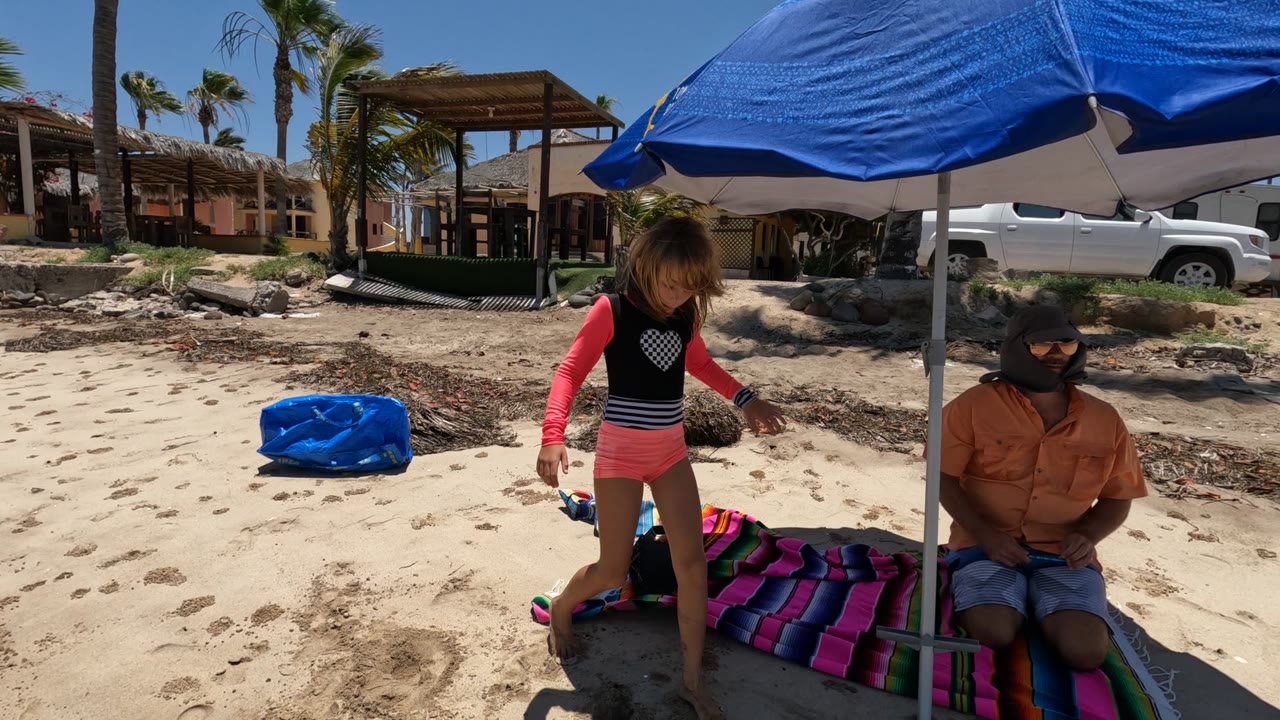 In Puerto San Carlos, Baja California, Mexico, a girl in pink shorts stands under a blue beach umbrella beside a seated adult on a colorful blanket, with palms and beach buildings behind them.