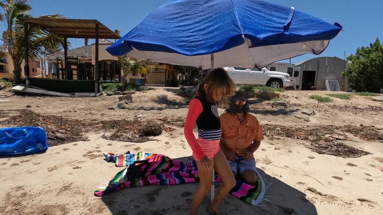 Girl stepping near a seated adult beneath a blue umbrella on a sandy beach.