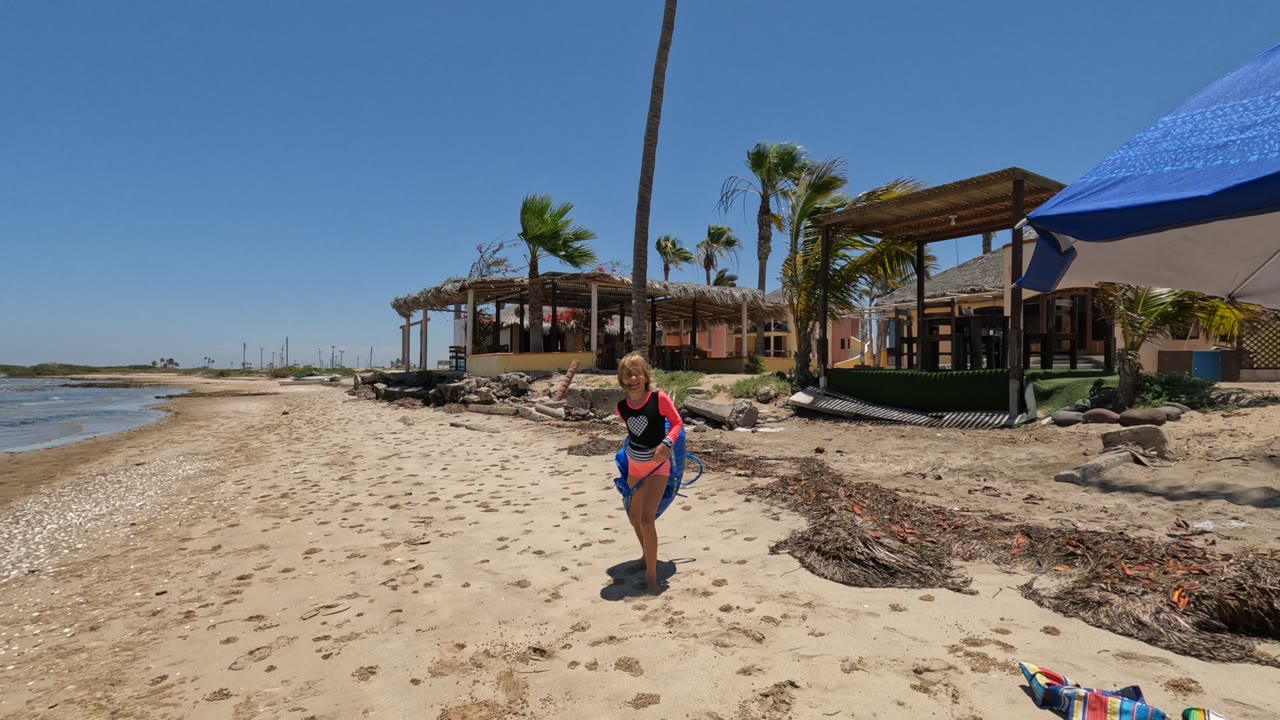 At 0:00 in Baja California, Mexico, a girl stands on the sandy beach near the waterline with a blue bag nearby, palm trees, a thatched structure, rocks, a blue umbrella, and a striped chair visible around her.