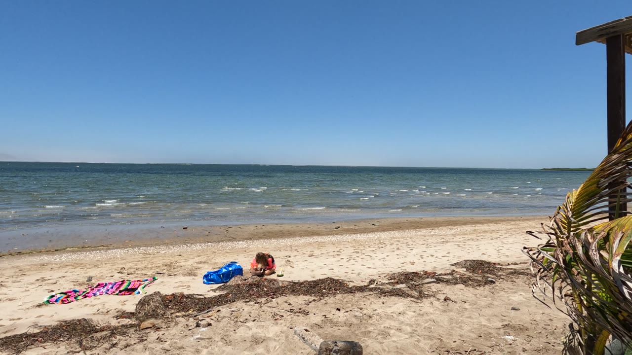 Baja California, Mexico beach view with a person on the sand near a blue towel, a striped towel at left, low ocean waves, and clear blue sky.