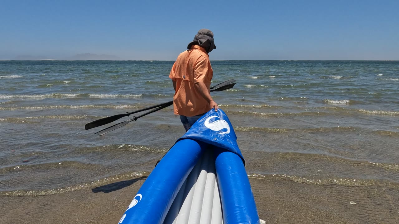 At 0:00 in Baja California, Mexico, a person in an orange shirt and hat stands near the front of a blue inflatable boat while holding paddles at the shoreline.