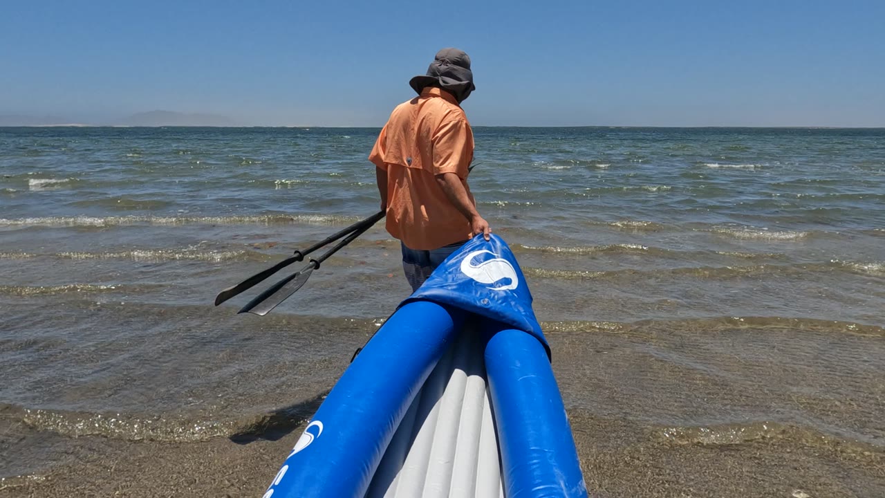 Person faces the ocean while holding a blue inflatable boat and paddles.