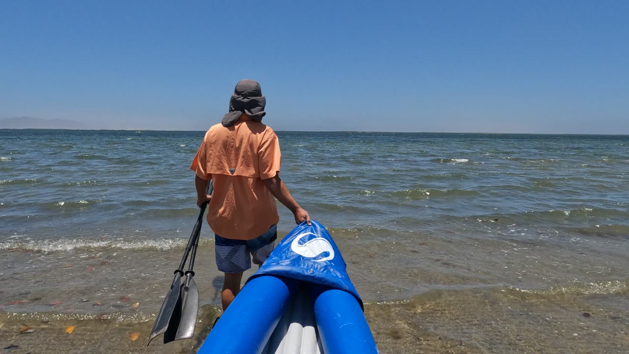 In Baja California, Mexico, a person in an orange shirt stands with a blue inflatable boat and paddle at the edge of shallow ocean water.