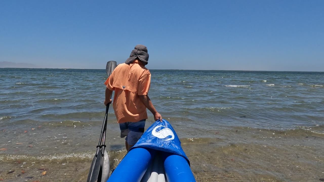 Person standing beside a blue inflatable boat with a paddle in shallow water.