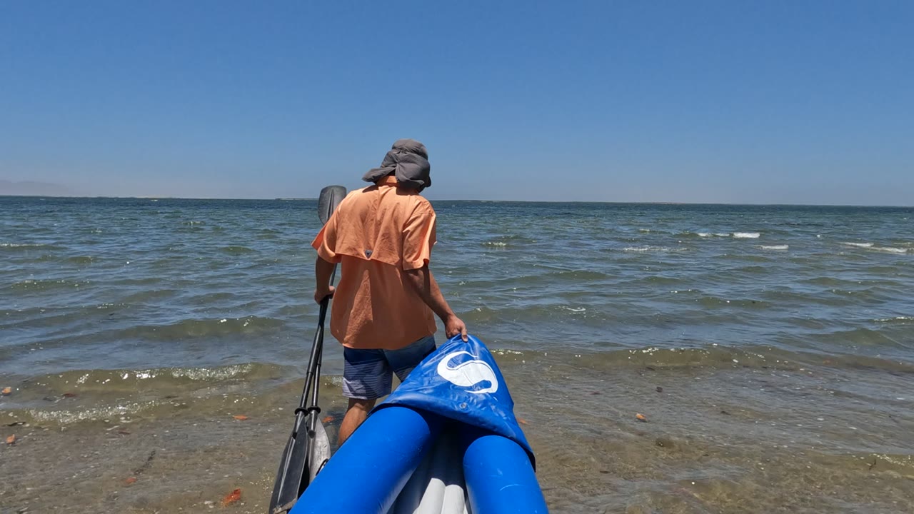 In Baja California, Mexico, the person stands near the right side of the frame with the blue inflatable boat angled into the shallow ocean.