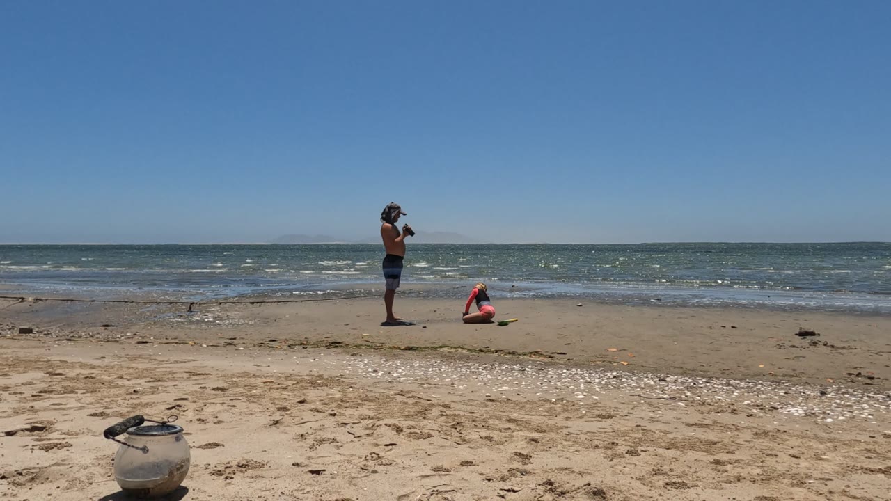 In Mulege, Mexico, one person stands with a camera while another crouches near the shoreline, with a pale container in the sandy foreground and blue water behind them.