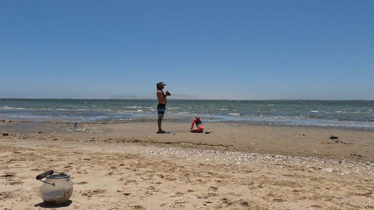 Standing person with camera and crouching person on beach by the ocean.