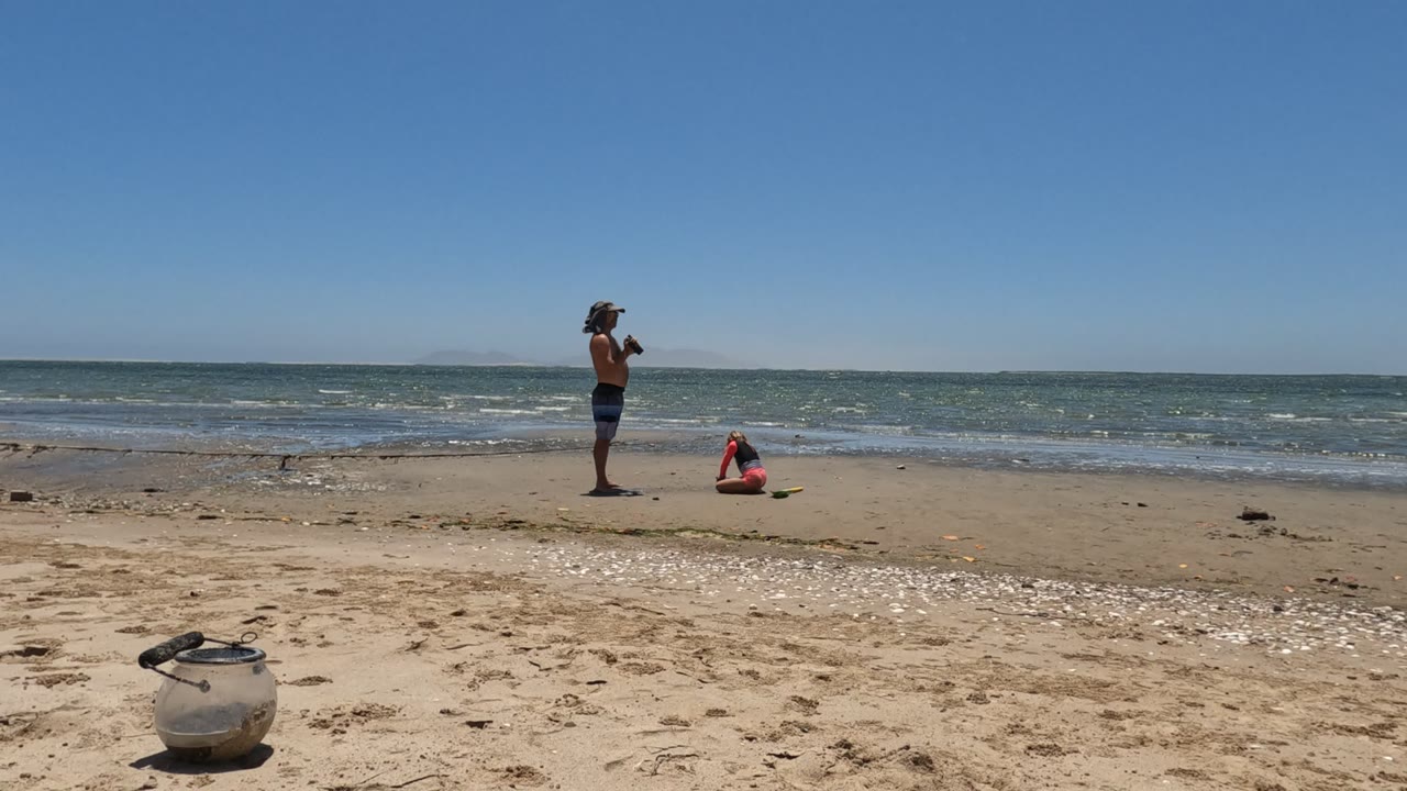 In Mulege, Mexico, the standing camera user and crouching beachgoer are spaced across the midground, with bright sand in front and a clear horizon over the ocean.