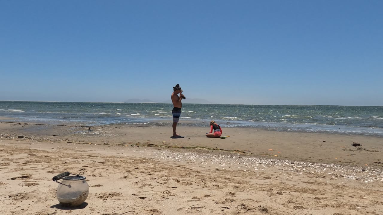 At 0:00 in Mulege, Baja California Mexico, one person stands near the surf while another sits on the sand, with a white bag in the foreground and low waves behind them.