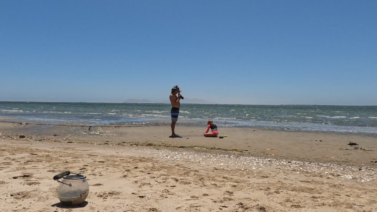 Standing person and seated person near small ocean waves on a sandy beach.