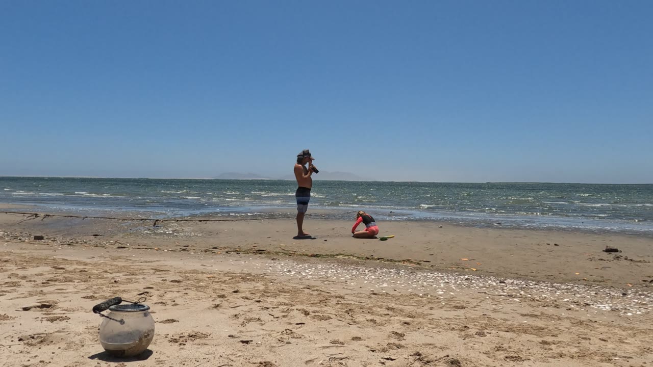 At 0:02 in Mulege, Baja California Mexico, two people remain separated on the open sand near the water, with bright shell scatter across the foreground and a flat blue horizon beyond the waves.