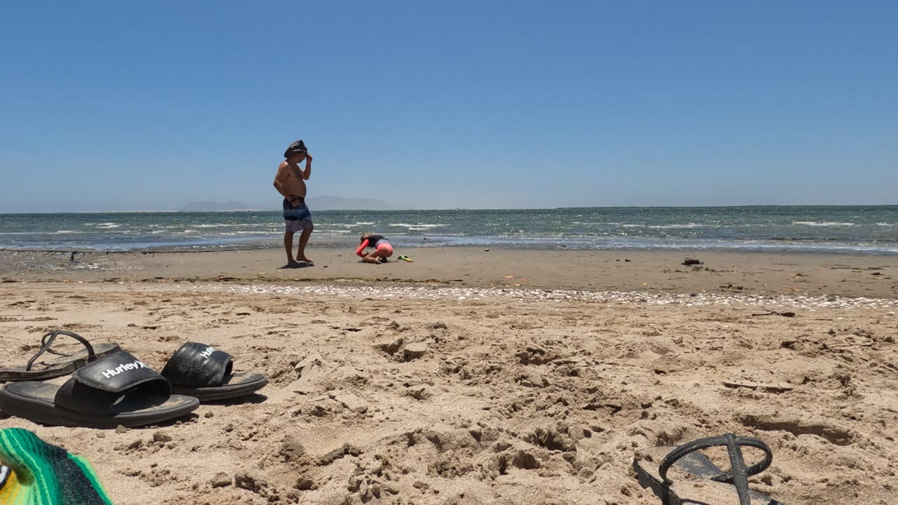In Baja California, Mexico, a shirtless man stands near the surf while a child in pink crouches by the water, with black flip-flops and a green towel on the sand in front.