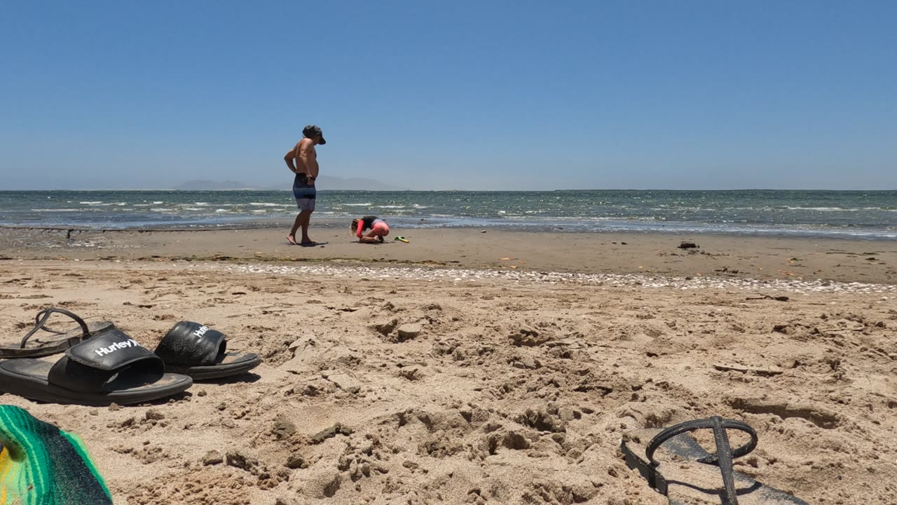 Man adjusting his hat while a child plays near the shoreline.