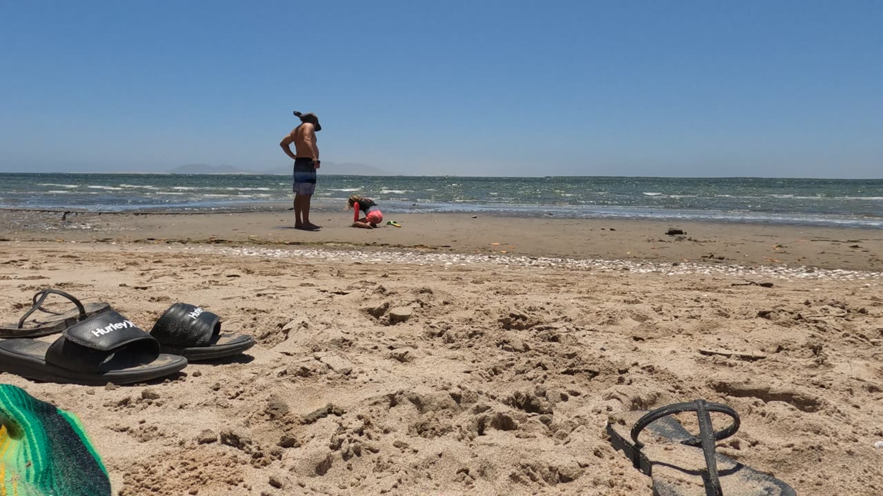 In Baja California, Mexico, the man stands upright near the waterline as the child crouches beside the surf, with the black flip-flops shifted toward the lower foreground of the frame.
