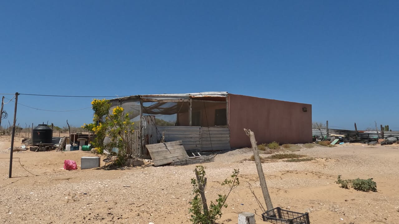 Wide view of a weathered shack in Baja California, Mexico, with sandy ground, rough fencing, a black water tank, scrub plants, and no visible people.