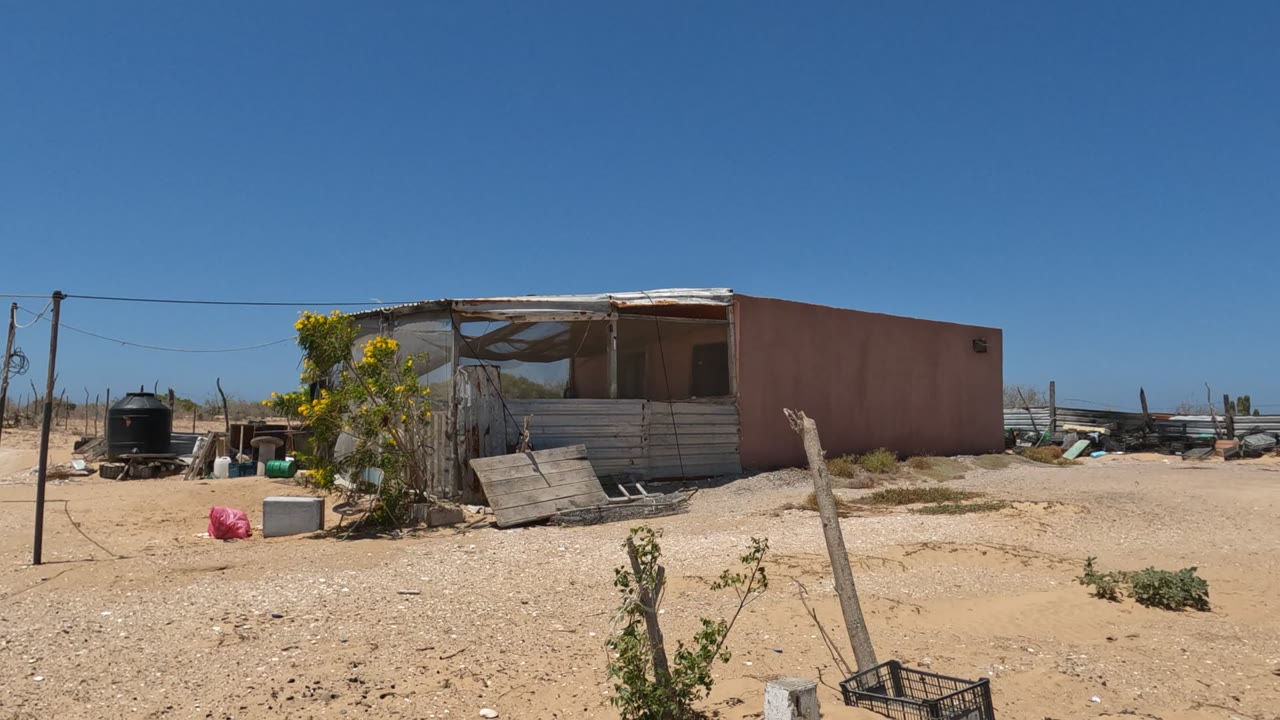 Desert shack yard with scattered containers and rough fencing.