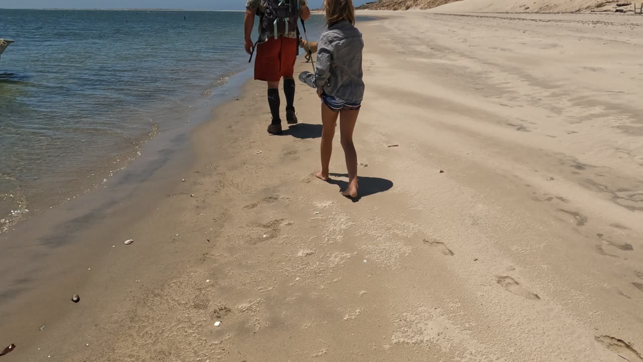A person in a denim jacket walks barefoot on the sandy Baja California, Mexico shoreline behind a backpacked beachgoer, with the small boat and calm water in the background.