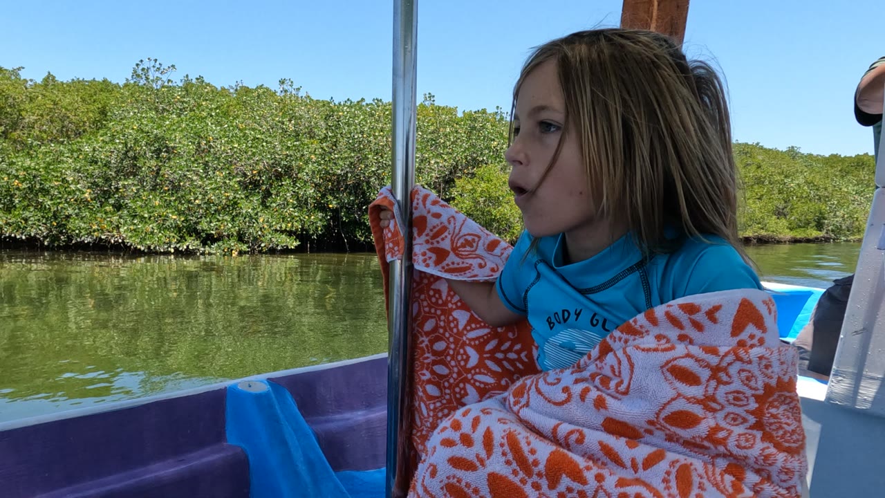A girl in Baja California, Mexico sits in profile on a boat, wrapped in an orange and white patterned blanket with green water and dense riverbank vegetation behind her.