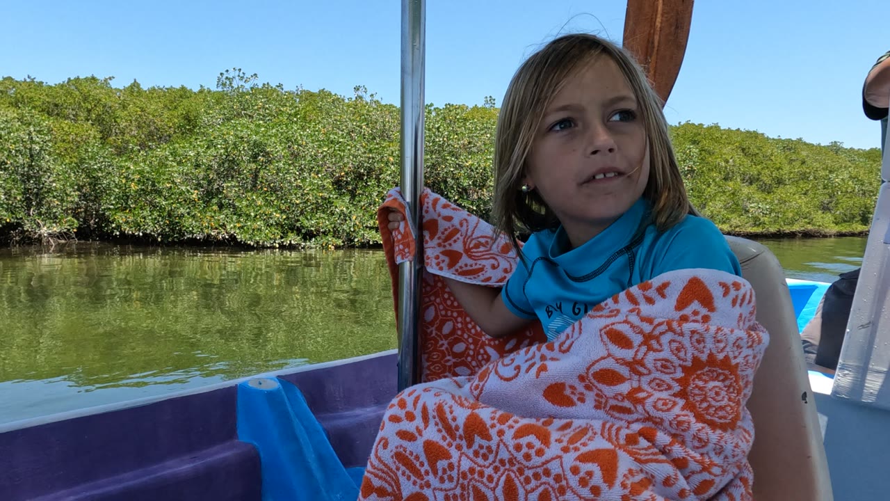 Girl on a boat turning toward the camera with green water and shrubs behind her.