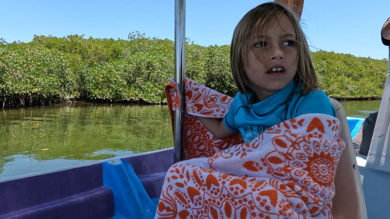 A girl in Baja California, Mexico faces forward in the boat with windblown hair, an orange patterned blanket across her lap, and sunlit green vegetation across the water.