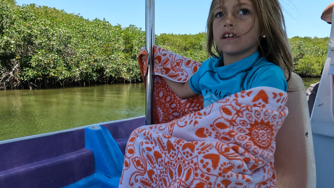 At the start of the Baja California, Mexico clip, a young girl in a blue shirt sits wrapped in an orange and white blanket beside calm green water and dense mangroves.